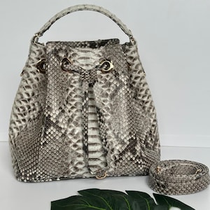 May include: A bucket-style handbag with a snakeskin pattern in shades of gray and white. The bag has a top handle and a detachable shoulder strap. Gold-colored hardware accents the bag's design. The bag is displayed on a white surface.