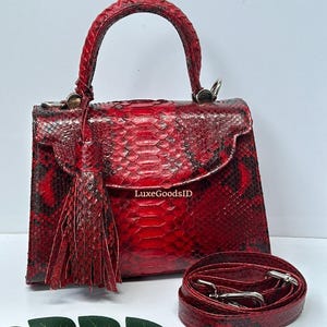 May include: Red snakeskin handbag with a top handle and detachable shoulder strap. The bag features a flap closure and a decorative tassel. The bag's exterior is a vibrant red with black accents, and the strap is coiled beside the bag.