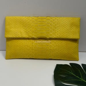May include: A bright yellow clutch with a textured, snakeskin-like pattern. The rectangular bag has a fold-over flap and is made of a soft material. The text "LuxeGoodsID" is visible on the bag.