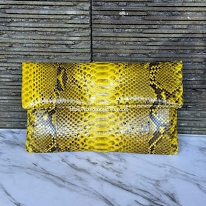 May include: A rectangular clutch bag featuring a vibrant yellow and black snakeskin pattern with a fold-over flap design. The surface displays a detailed scale texture throughout the exterior. The text https://luxegoodsid.etsy.com is visible.
