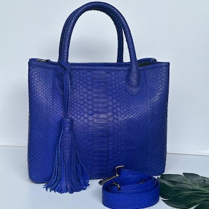 Könnte beinhalten: Eine kobaltblaue Tragetasche mit einem strukturierten Schlangenmuster. Die Tasche hat zwei obere Griffe, einen abnehmbaren Schultergurt und eine Quaste. Die Tasche ist etwa 30 cm hoch und 25 cm breit.