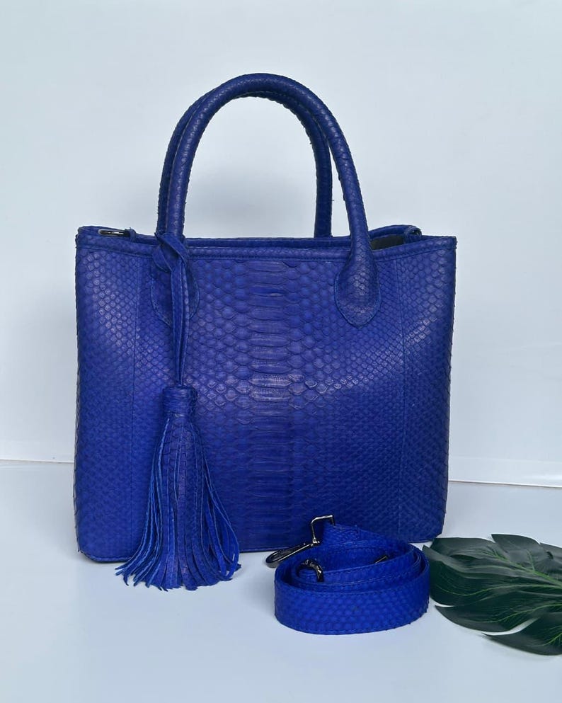 May include: A cobalt blue tote bag with a textured snakeskin pattern. The bag has two top handles, a detachable shoulder strap, and a tassel. The bag is approximately 12 inches tall and 10 inches wide.