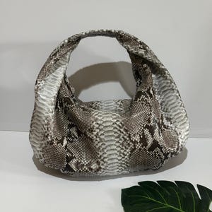 Könnte beinhalten: Eine große, lässige Handtasche mit Schlangenmuster in Grau-, Braun- und Weißtönen. Die Tasche hat eine runde Form mit einem einzelnen, breiten Griff. Die Tasche ist aus einem strukturierten Material gefertigt.