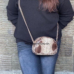 Peut inclure: Petit sac &agrave; main ovale avec un motif peau de serpent marron et cr&egrave;me. Il est dot&eacute; d'une bandouli&egrave;re dor&eacute;e et d'une fermeture &eacute;clair. Le sac est port&eacute; &agrave; l'&eacute;paule. Dimensions: environ 20 cm de long.