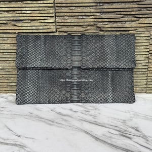 May include: A rectangular black clutch bag featuring a textured snakeskin pattern with a prominent central spine detail running vertically down the middle. The surface displays a consistent scale texture throughout, and the text https://luxegoodsid.etsy.com is printed in the center.