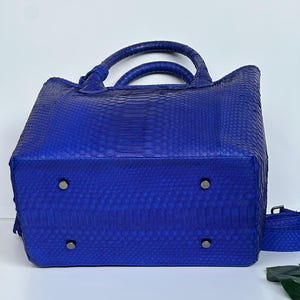 May include: A cobalt blue handbag with a snakeskin pattern. The bag has two rounded handles and a detachable shoulder strap. The bottom of the bag has four metal feet.