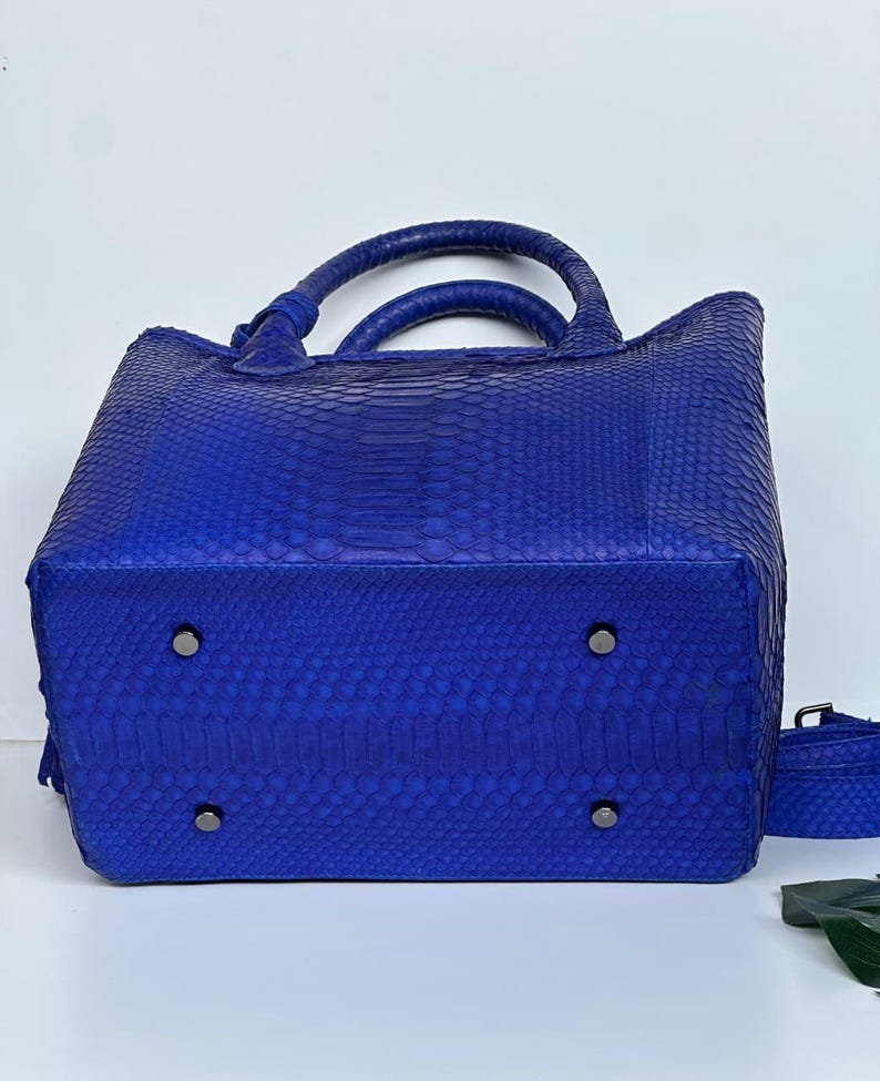 May include: A cobalt blue handbag with a snakeskin pattern. The bag has two rounded handles and a detachable shoulder strap. The bottom of the bag has four metal feet.