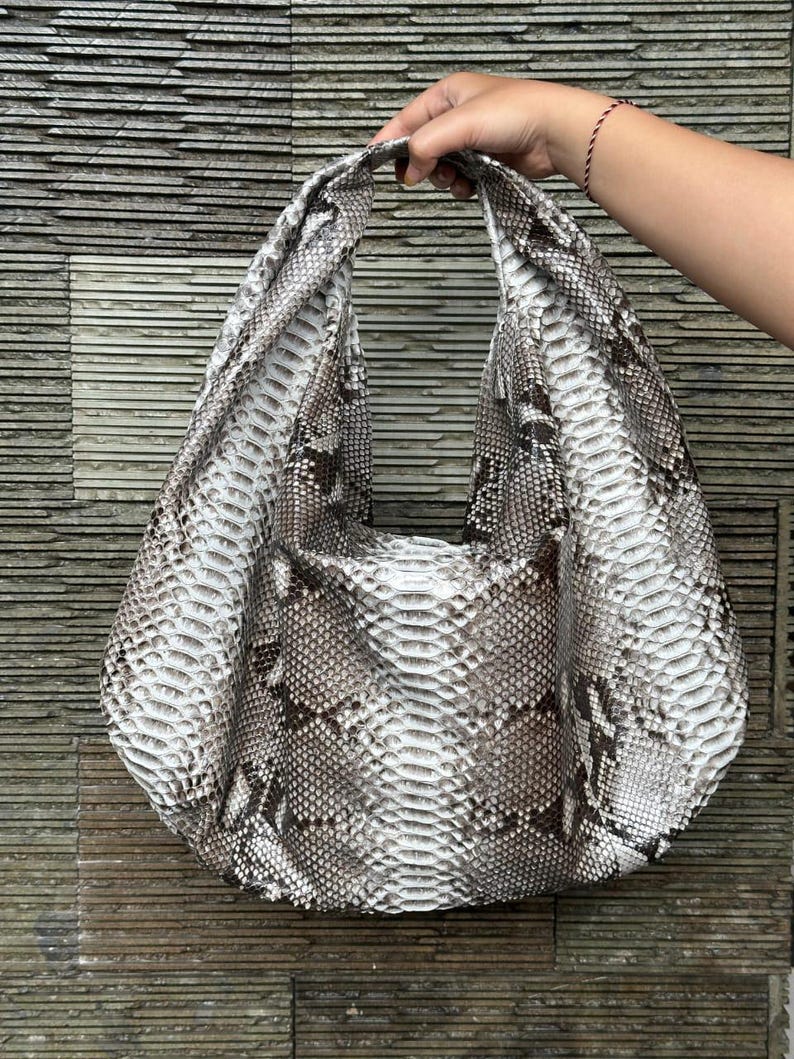 May include: A large, open-top handbag with a snakeskin pattern in shades of white, gray, and brown. The bag has a single, wide handle and a rounded shape. The bag is held up by a hand.