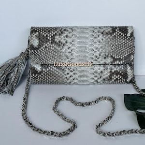Peut inclure: Pochette à motif peau de serpent avec une bandoulière chaîne argentée et un pompon assorti. Le sac est doté d'une fermeture à rabat et du texte "LuxeGoodsID" en lettres dorées. La palette de couleurs comprend des nuances de gris, de blanc et de noir.