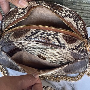 Peut inclure: Un sac &agrave; main rond avec un motif peau de serpent et une cha&icirc;ne dor&eacute;e. Il est dot&eacute; d'une fermeture &eacute;clair marron et d'une petite poche. L'int&eacute;rieur est de couleur marron clair. Le sac est ouvert, r&eacute;v&eacute;lant l'int&eacute;rieur.