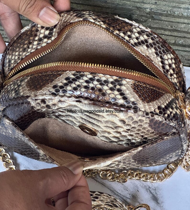 Peut inclure: Un sac &agrave; main rond avec un motif peau de serpent et une cha&icirc;ne dor&eacute;e. Il est dot&eacute; d'une fermeture &eacute;clair marron et d'une petite poche. L'int&eacute;rieur est de couleur marron clair. Le sac est ouvert, r&eacute;v&eacute;lant l'int&eacute;rieur.