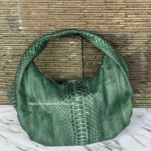 May include: A green, textured handbag with a rounded handle. The bag has a snakeskin pattern and a curved shape. The Etsy shop URL is visible on the bag. The bag is made of a material that resembles snakeskin.