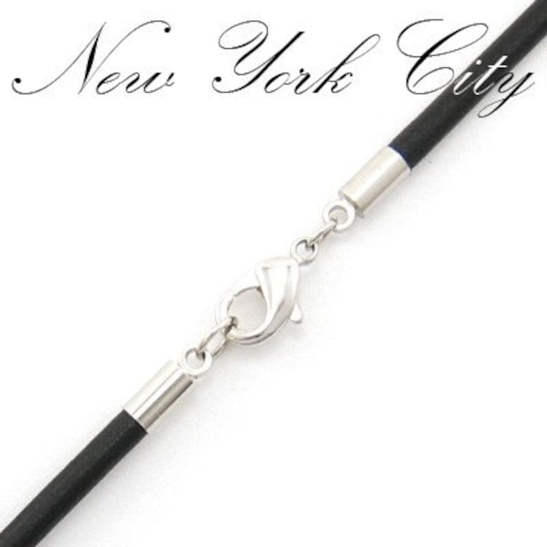 May include: Close-up of a black cord necklace with a silver-colored clasp and accents. The words "New York City" are written in a cursive font at the top of the image.