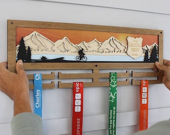 May include: A wooden medal display rack with a mountain scene and a cyclist silhouette. The rack has slots for hanging medals and ribbons. The ribbons are red, green, and blue with text that includes "AQUASPHERE", "epic", "Chorley", and "UK Kids Fun Duathlon Cheshire".