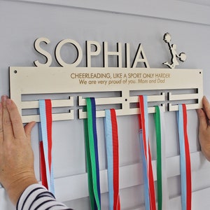 Può includere: Un porta medaglie in legno con il nome "SOPHIA" e il testo "CHEERLEADING, LIKE A SPORT ONLY HARDER We are very proud of you. Mom and Dad" inciso su di esso. Il porta medaglie ha cinque slot per le medaglie e una silhouette di una cheerleader con pompon.