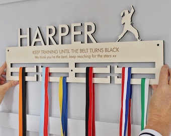 Könnte beinhalten: Ein hölzernes Medaillendisplay mit dem ausgeschnittenen Namen "HARPER" oben. Darunter steht der Text "KEEP TRAINING UNTIL THE BELT TURNS BLACK". Bunte Bänder hängen an der Anzeige. Eine Silhouette eines Kampfsportlers befindet sich auf der rechten Seite.