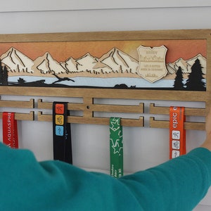 Op de afbeelding: Een houten medaillescherm met een berglandschap en een gepersonaliseerd bord met de tekst "Daddy, Life is better when you're camping". Het scherm heeft vier vakken voor medailles en wordt vastgehouden door de handen van een persoon.