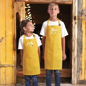 May include: Two children wearing yellow aprons with their names, Alex and Millie, embroidered on them. The aprons have adjustable straps and a pocket. The children are standing in front of a wooden door.