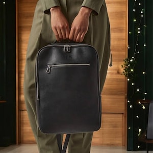 May include: A black leather backpack with silver hardware and a zippered pocket. The backpack has a small silver tag with the letters "TRS" on it.