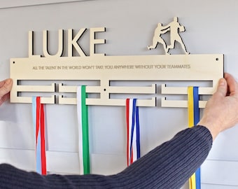 Könnte beinhalten: Medaillenhalter aus Holz mit dem ausgeschnittenen Namen "LUKE" oben. Die Halterung hat Schlitze für Medaillen mit bunten Bändern. Der Text "ALL THE TALENT IN THE WORLD WON'T TAKE YOU ANYWHERE WITHOUT YOUR TEAMMATES" ist eingraviert.