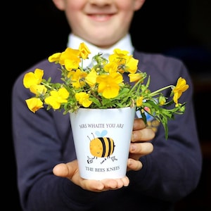 May include: A child is holding a white ceramic mug with a yellow and black bee illustration and the text "Mrs. Whaite you are the bees knees". The mug is filled with yellow flowers.