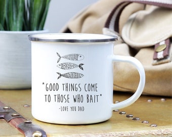 May include: White enamel mug with a silver rim and a handle. The mug features three illustrated fish and the text "GOOD THINGS COME TO THOSE WHO BAIT" and "-LOVE YOU DAD" in black. The mug is on a wooden surface.