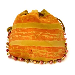 May include: A drawstring pouch with a striped orange and yellow pattern. The pouch is trimmed with a border of pink and gold beads. The top of the pouch is gathered with a green drawstring.