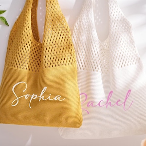 May include: Two knitted tote bags, one mustard yellow and one white, hang against a white background. The yellow bag has the name "Sophia" in white script, and the white bag has the name "Rachel" in pink script. The bags have a mesh-like design.