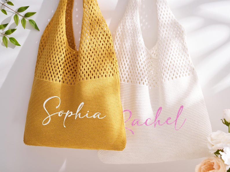 May include: Two knitted tote bags, one mustard yellow and one white, hang against a white background. The yellow bag has the name "Sophia" in white script, and the white bag has the name "Rachel" in pink script. The bags have a mesh-like design.