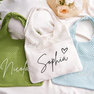 May include: Three personalised tote bags in green, white, and light blue. The bags have a knitted mesh upper section and a solid lower section with a name embroidered on each. The white bag has a heart symbol next to the name.