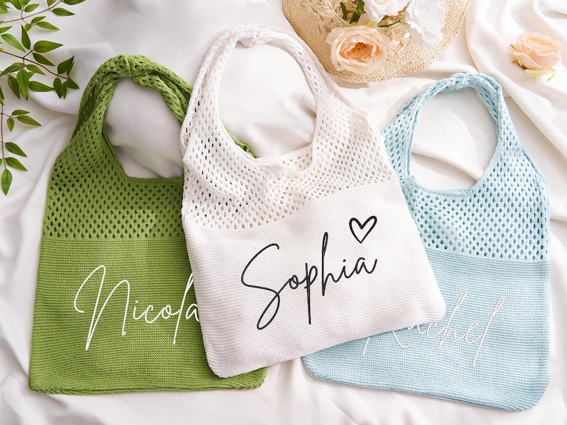 May include: Three personalised tote bags in green, white, and light blue. The bags have a knitted mesh upper section and a solid lower section with a name embroidered on each. The white bag has a heart symbol next to the name.