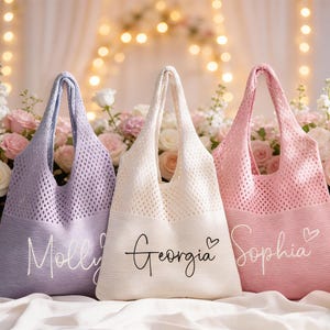 May include: Three pastel-coloured tote bags: lavender, cream, and pink. Each bag features a knitted mesh design and a solid bottom with a name in script. The bags are displayed with a floral arrangement.