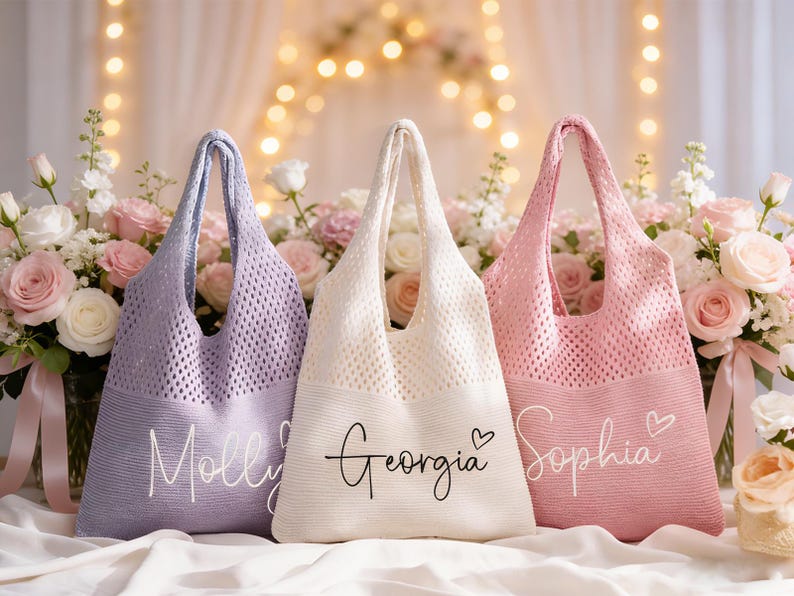 May include: Three pastel-coloured tote bags: lavender, cream, and pink. Each bag features a knitted mesh design and a solid bottom with a name in script. The bags are displayed with a floral arrangement.