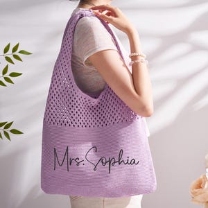 May include: A lilac crocheted tote bag with the text "Mrs. Sophia" in black script. The bag features an open weave at the top and a solid knit base. The bag is being held by a person.