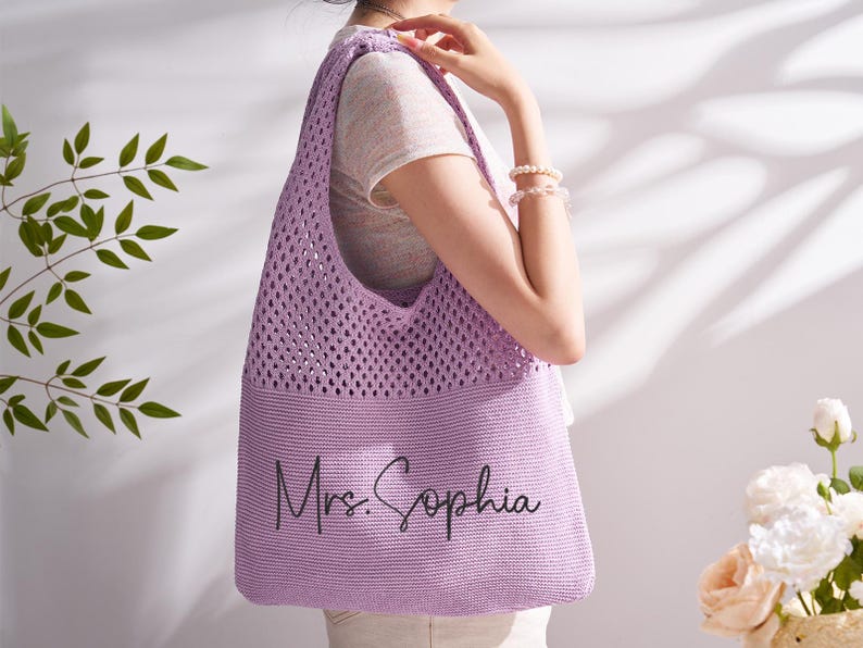 May include: A lilac crocheted tote bag with the text "Mrs. Sophia" in black script. The bag features an open weave at the top and a solid knit base. The bag is being held by a person.