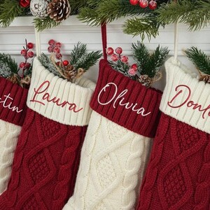 May include: Four personalized Christmas stockings. Each stocking is cream-colored with a red cuff and cable knit design. The names "Justin", "Laura", "Olivia", and "Donlon" are embroidered in white script. Greenery and berries adorn the top.