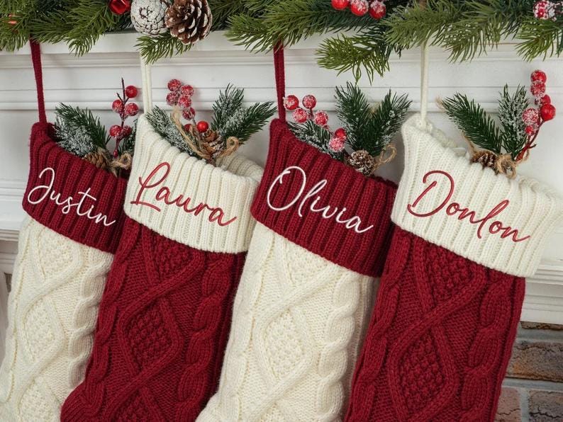 May include: Four personalized Christmas stockings. Each stocking is cream-colored with a red cuff and cable knit design. The names "Justin", "Laura", "Olivia", and "Donlon" are embroidered in white script. Greenery and berries adorn the top.