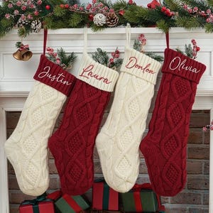 May include: Four cable-knit Christmas stockings hang from a decorated mantel. Two are cream-colored, and two are red. Each stocking has a name embroidered on the cuff: Justin, Laura, Donlon, and Olivia. The mantel is adorned with greenery and berries.