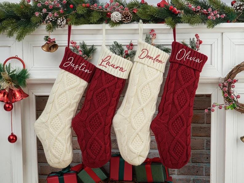 May include: Four cable-knit Christmas stockings hang from a decorated mantel. Two are cream-colored, and two are red. Each stocking has a name embroidered on the cuff: Justin, Laura, Donlon, and Olivia. The mantel is adorned with greenery and berries.