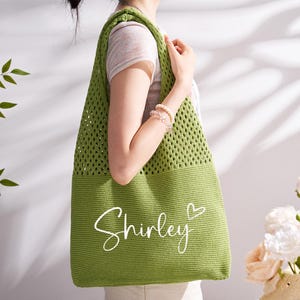 May include: A green crochet tote bag with the name "Shirley" in white script and a heart symbol. The bag has a mesh design on the upper portion and a solid bottom. The bag is being held by a person.