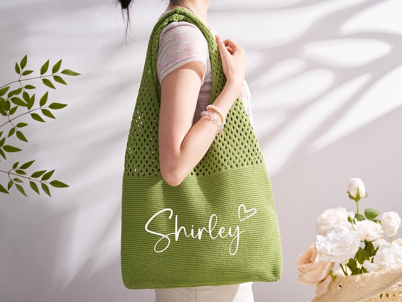 May include: A green crochet tote bag with the name "Shirley" in white script and a heart symbol. The bag has a mesh design on the upper portion and a solid bottom. The bag is being held by a person.