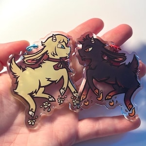 Op de afbeelding: Twee acryl geitenbedels, één crèmekleurig en één zwart, worden in een hand gehouden. De crèmekleurige geit heeft blauwe ogen en een rode strik, terwijl de zwarte geit rode ogen heeft. Beide hebben bloemmotieven.