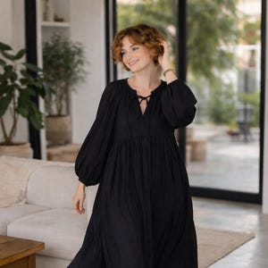 May include: A long, black dress with long, voluminous sleeves and a tie-neck detail. The dress is flowing and appears to be made of a lightweight fabric. The model is wearing brown sandals. The setting is a well-lit room with a neutral color palette.