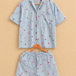 May include: A light blue and white striped pajama set with pink heart accents. The set includes a short-sleeved button-up shirt and matching shorts. The shirt has a chest pocket and the shorts have an elastic waistband.