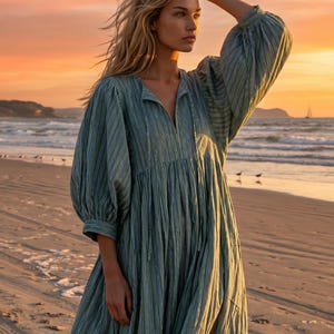 May include: A long, flowing dress with vertical stripes in shades of green and white. The dress features a V-neckline and voluminous sleeves. The photo is taken on a beach at sunset, with the ocean and sky in the background.