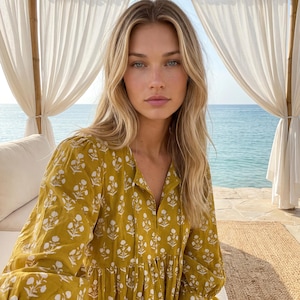 May include: A mustard yellow long-sleeved dress featuring a repeating white floral block print pattern, a split V-neckline with a tie closure, and a gathered waistline that creates a loose, flowing silhouette with cuffed sleeves.
