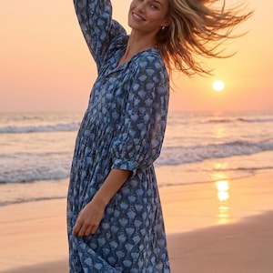 May include: A blue floral print dress with long sleeves and a gathered waist. The dress is worn on a beach at sunset. The model is smiling and has her arm raised. The dress is knee-length.