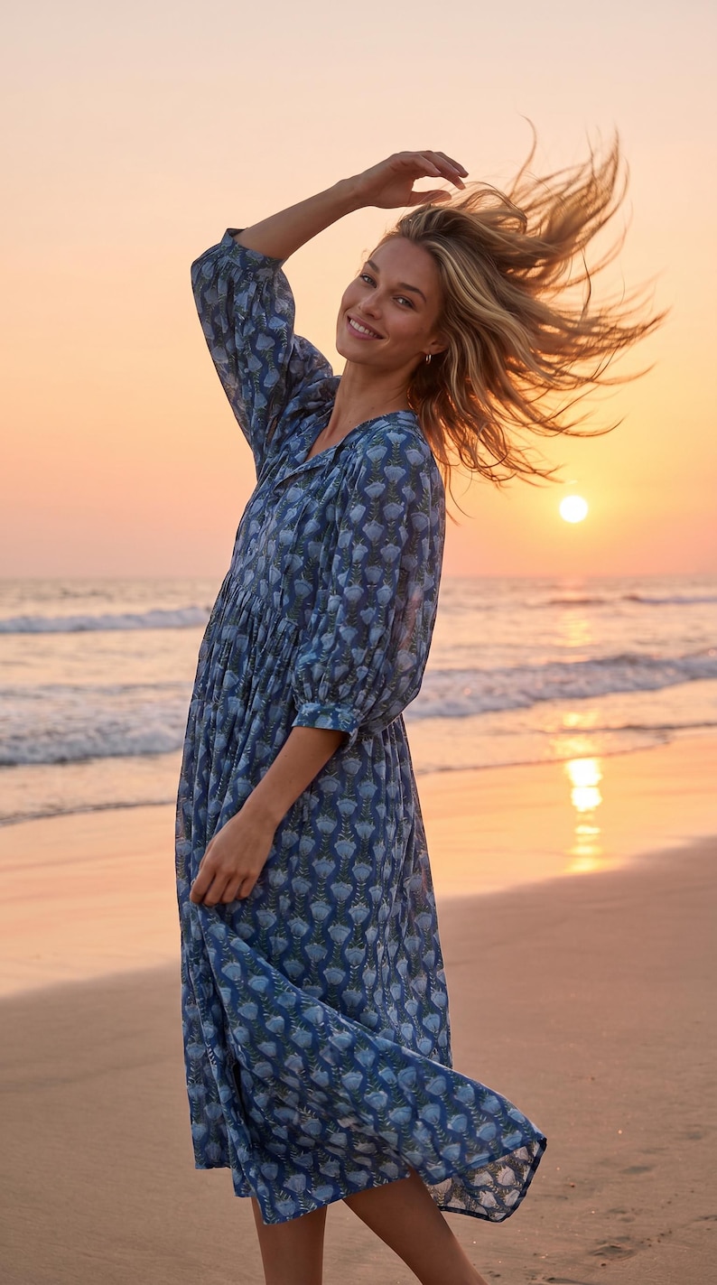 May include: A blue floral print dress with long sleeves and a gathered waist. The dress is worn on a beach at sunset. The model is smiling and has her arm raised. The dress is knee-length.