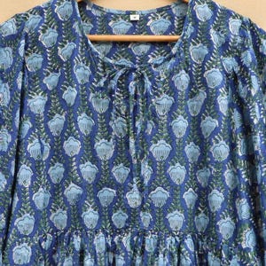 May include: A blue dress with a floral design. The dress features a round neckline with a tie detail and long sleeves. The floral pattern is light blue and green on a dark blue backdrop. The dress is displayed on a wooden hanger.