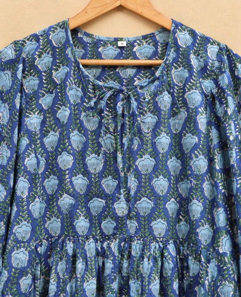 May include: A blue dress with a floral design. The dress features a round neckline with a tie detail and long sleeves. The floral pattern is light blue and green on a dark blue backdrop. The dress is displayed on a wooden hanger.